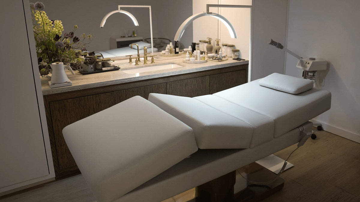 goop's first spa treatment room at Marin Country Mart: an Oxford electric salon bed in Bone on a bronzed brass base, beneath an arched mirror alcove with a marble counter and an LED arc lamp.