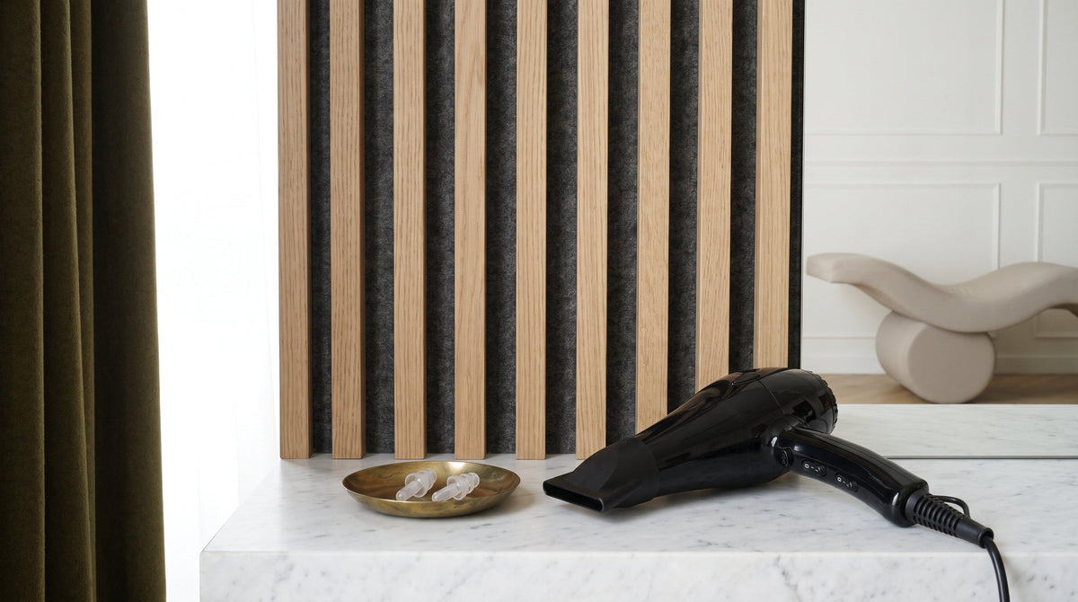 Filtered musicians' earplugs in a brass dish beside a hair dryer on a marble ledge, against a fluted white oak wall with grey acoustic felt between the slats.