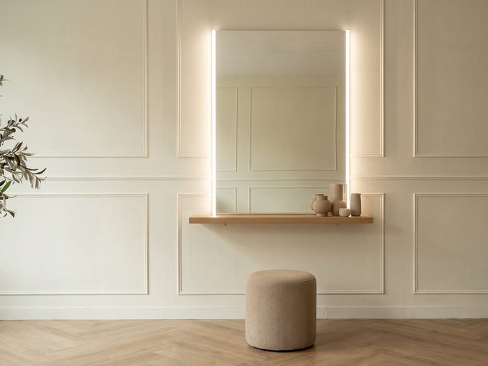 A salon styling station lit by slim vertical LED strips running down both sides of a tall mirror at face height, above a slim oak console shelf and upholstered stool.