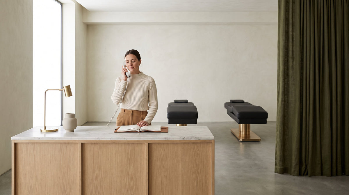 A salon receptionist takes a phone call at a white oak reception desk with an open appointment book, two charcoal Oxford electric salon beds on brass bases on the treatment floor behind her.