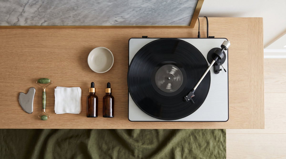 Overhead view of a turntable with a record playing on a white oak salon shelf, a gua sha, jade roller, folded gauze and amber dropper bottles laid out beside it.