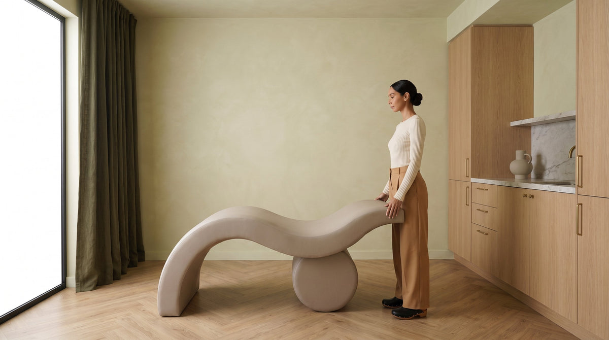 A lash artist in a cream top and camel trousers stands beside a Brynn curved salon bed on its cylindrical drum base, in a treatment room with white oak joinery, a marble counter and a deep olive curtain.