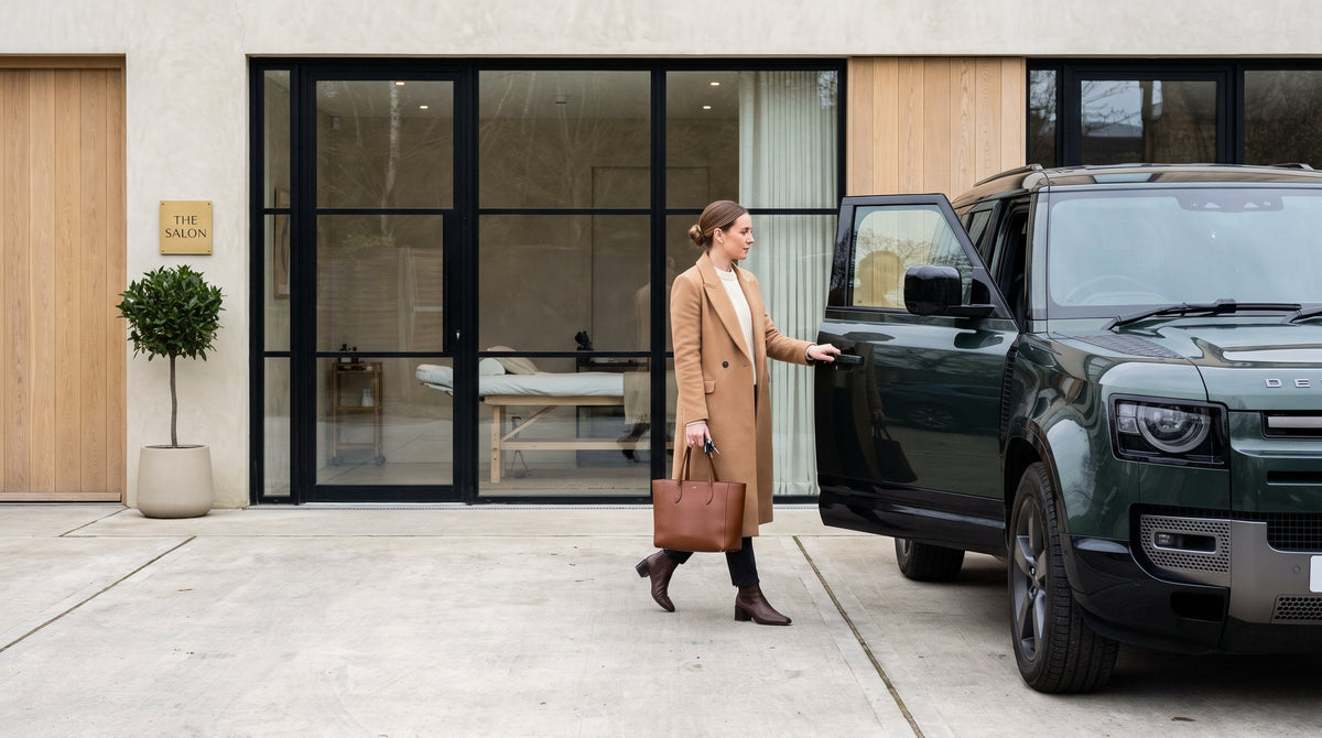 A businesswoman in a camel coat leaves a salon and opens the door of a dark green SUV in the car park, the treatment room visible through the glass frontage behind her.